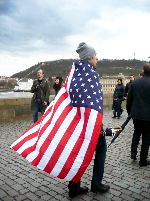 Muž zahalený v americké vlajce prochází přes Karlův most.